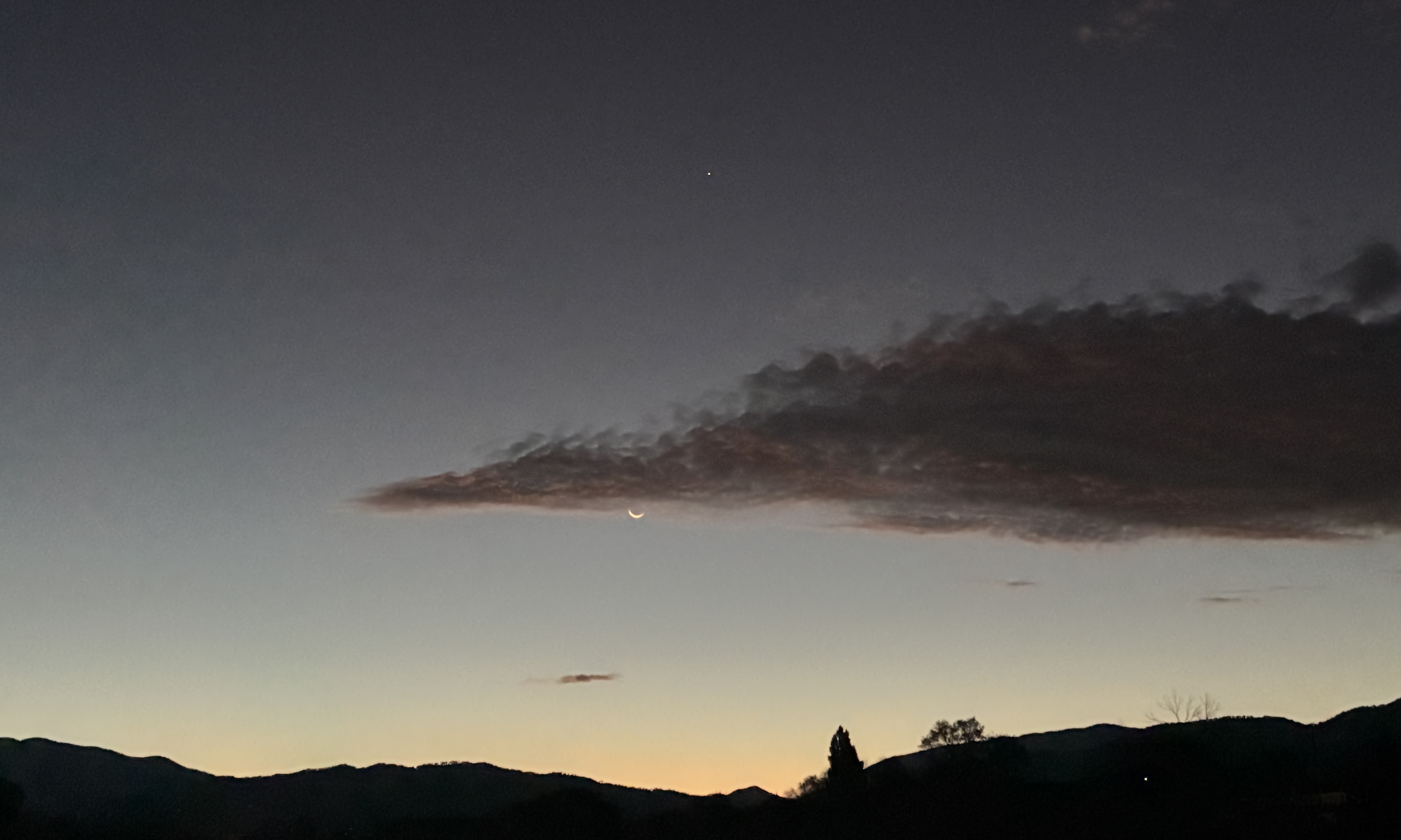 photo of sunrise in New Mexico featuring a hilly horizon with a sky fading almost to black from the violet hint of the rising sun
the center of the photo is a dark cloud emerging in a point from the right with the crescent moon seen just below and Venus clear above the cloud, almost directly above the moon